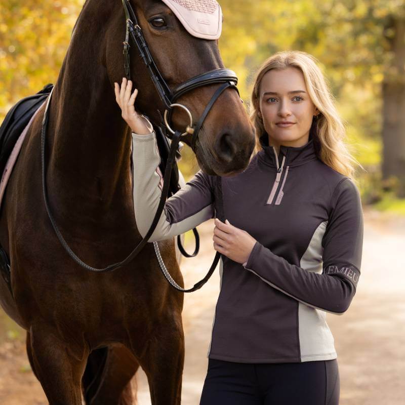 Woman in LeMieux Matila Midlayer Cocoa smiles by her horse, showing equestrian style.