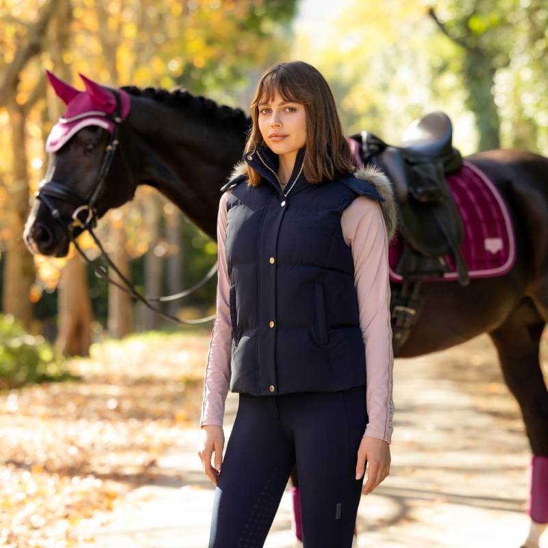 Woman in a LeMieux Margot Hooded Gilet Navy stands by a black horse with pink tack on a path.