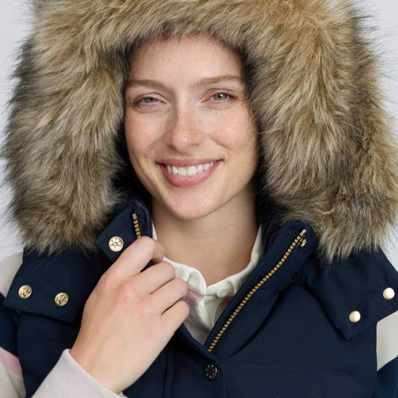Woman smiles in LeMieux Margot Hooded Gilet Navy, holding its fur-lined collar with one hand.