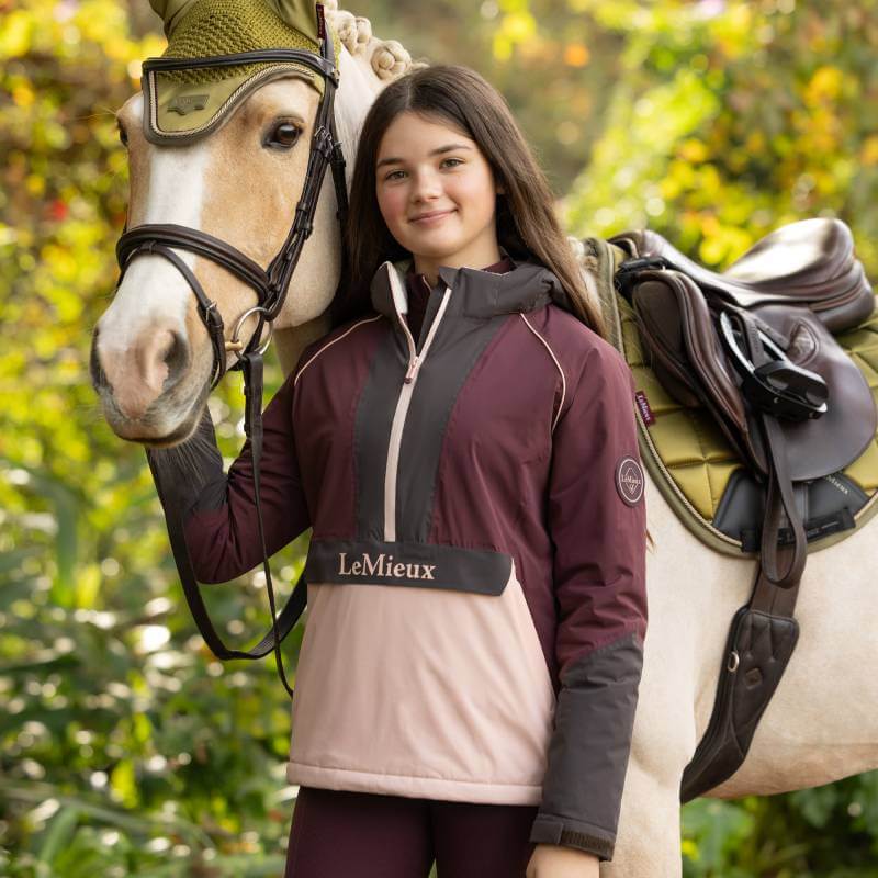 Girl in LeMieux Young Rider Bailey Anorak stands by a light horse with a green saddle.
