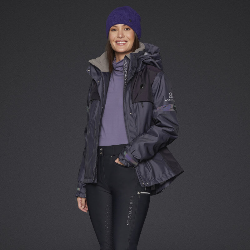 A woman wears a Mountain Horse Royal Grace Jacket Deep Purple, purple beanie, shirt, black pants.