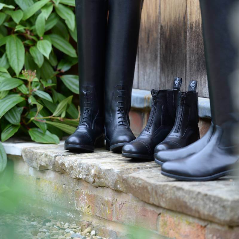Three pairs of Saxon Syntovia Tall Dress Boots lined up on a stone ledge by green plants.