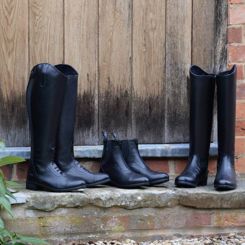 Three pairs of Saxon Syntovia Tall Dress Boots in black rest on a stone step by a door.