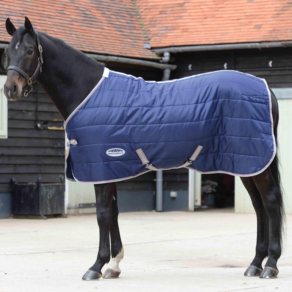 A black horse wears the Weatherbeeta Comfitec 210D Quilt rug in a stable yard with brick buildings.