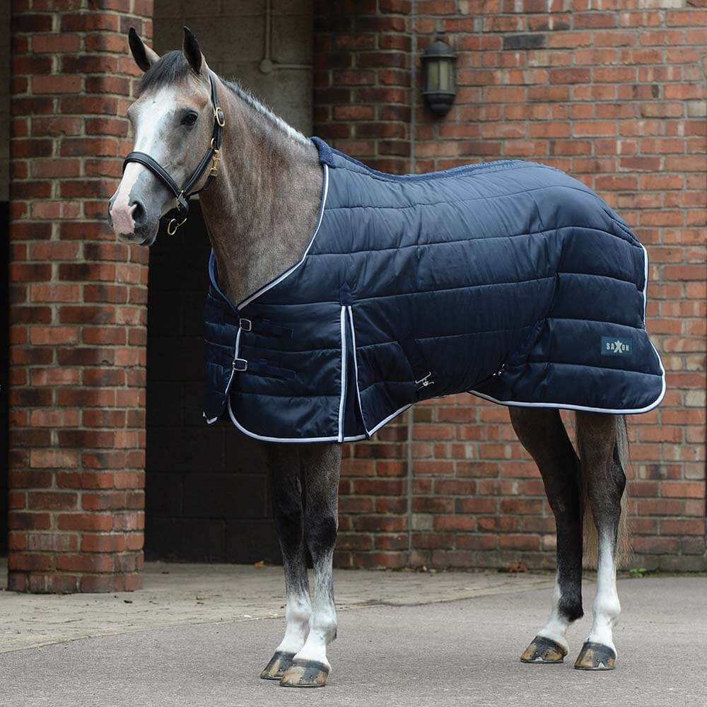 A horse in a Saxon Channel Quilt Stable Rug Standard Neck Navy/White stands by a brick wall.
