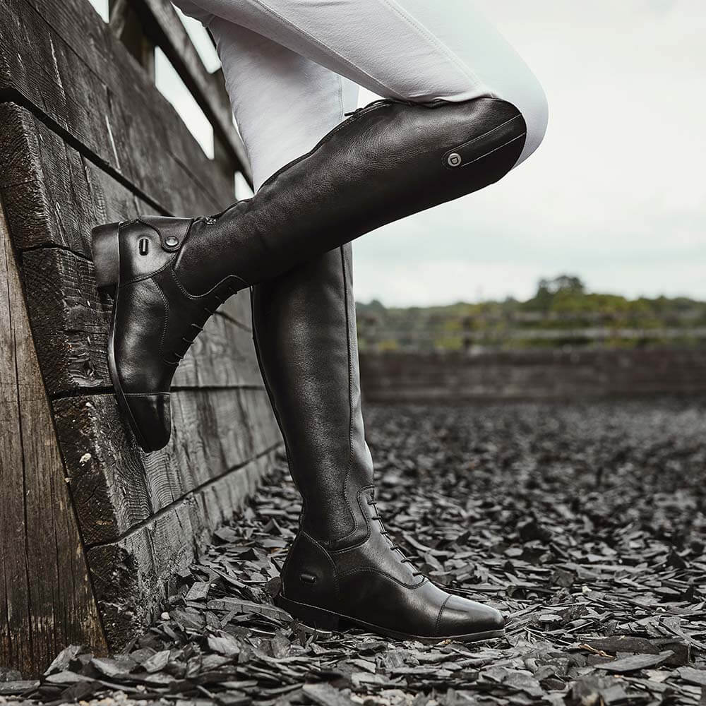 Person wearing Dublin Arderin Tall Field Boots, leaning on a wooden fence outdoors.