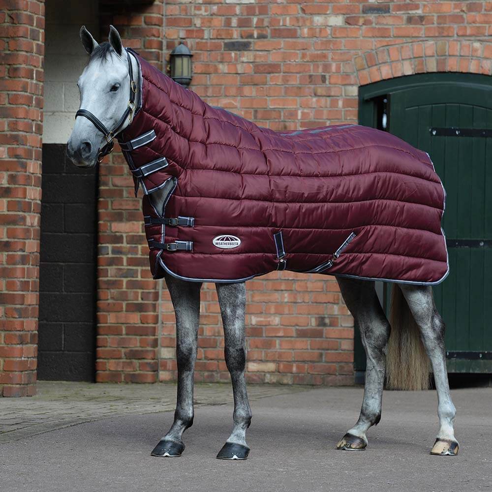 A horse wears the Weatherbeeta Comfitec 210D maroon combo neck rug by a brick stable.