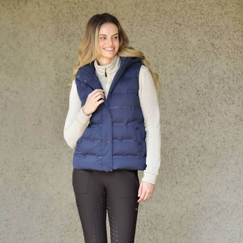 Woman wearing a Weatherbeeta Hapur Heat Seal Quilted Vest in Navy Iris, smiling by a textured wall.