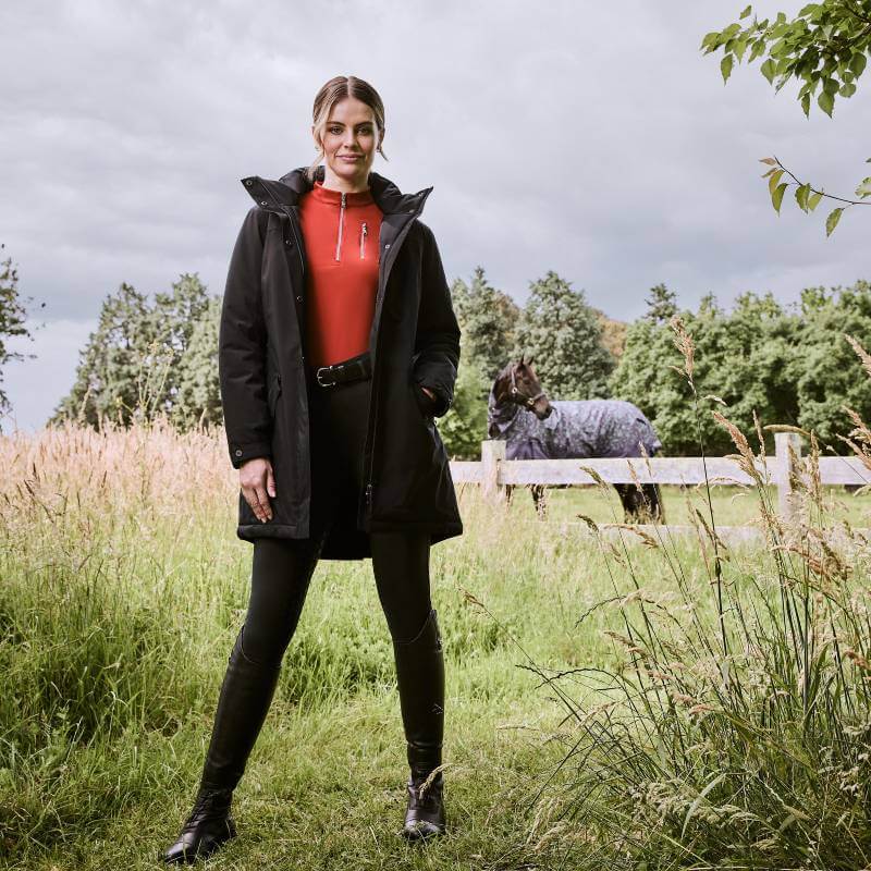 Woman wears a Weatherbeeta Kyla II Waterproof Jacket Black in a grassy field with a horse behind.