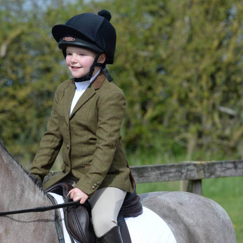 Child wears Dublin Albany Tweed Jacket and helmet while sitting on a horse outdoors.